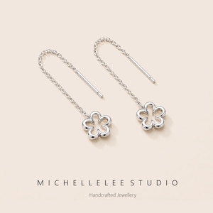 Peut inclure: Une paire de boucles d'oreilles argentées en forme de fleur. Chaque boucle d'oreille est ornée d'une chaîne délicate menant à un charm en forme de fleur. Le texte "MICHELLELEE STUDIO" et "Handcrafted Jewellery" est en bas.