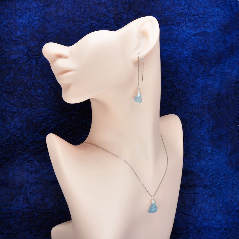 May include: A silver necklace and earrings set with light blue gemstone pendants. The necklace and earrings feature a delicate chain and a simple design.