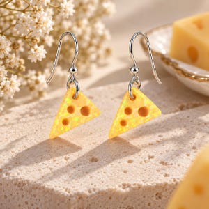 May include: A pair of drop earrings featuring triangular charms shaped like slices of yellow cheese with small circular cutouts. The charms have a textured, iridescent finish and are suspended from silver-toned metal hooks.