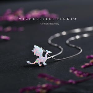 May include: A silver necklace with a dragon pendant. The dragon pendant is white with pink and green iridescent accents. The necklace is on a dark surface with the text "MICHELLELEE STUDIO Handcrafted Jewellery".
