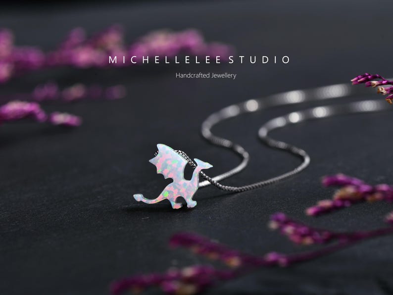May include: A silver necklace with a dragon pendant. The dragon pendant is white with pink and green iridescent accents. The necklace is on a dark surface with the text "MICHELLELEE STUDIO Handcrafted Jewellery".