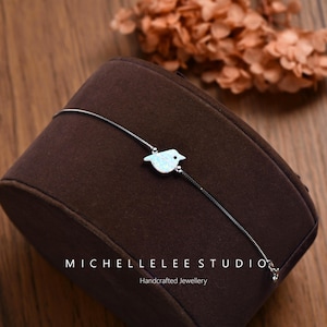 Peut inclure: Un bracelet en argent délicat orné d'un petit oiseau blanc. L'oiseau présente un léger éclat opalescent. Le bracelet est présenté sur une surface en velours marron. Le texte "MICHELLELEE STUDIO" et "Handcrafted Jewellery" sont visibles.