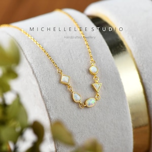 May include: A gold chain necklace with five white opal stones in different shapes, including a circle, a teardrop, a triangle, and a square. The necklace is on a white velvet background. The text "MICHELLELEESTUDIO Handcrafted Jewellery" is visible above the necklace.