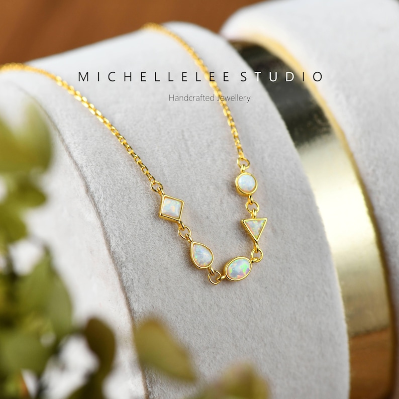 May include: A gold chain necklace with five white opal stones in different shapes, including a circle, a teardrop, a triangle, and a square. The necklace is on a white velvet background. The text "MICHELLELEESTUDIO Handcrafted Jewellery" is visible above the necklace.