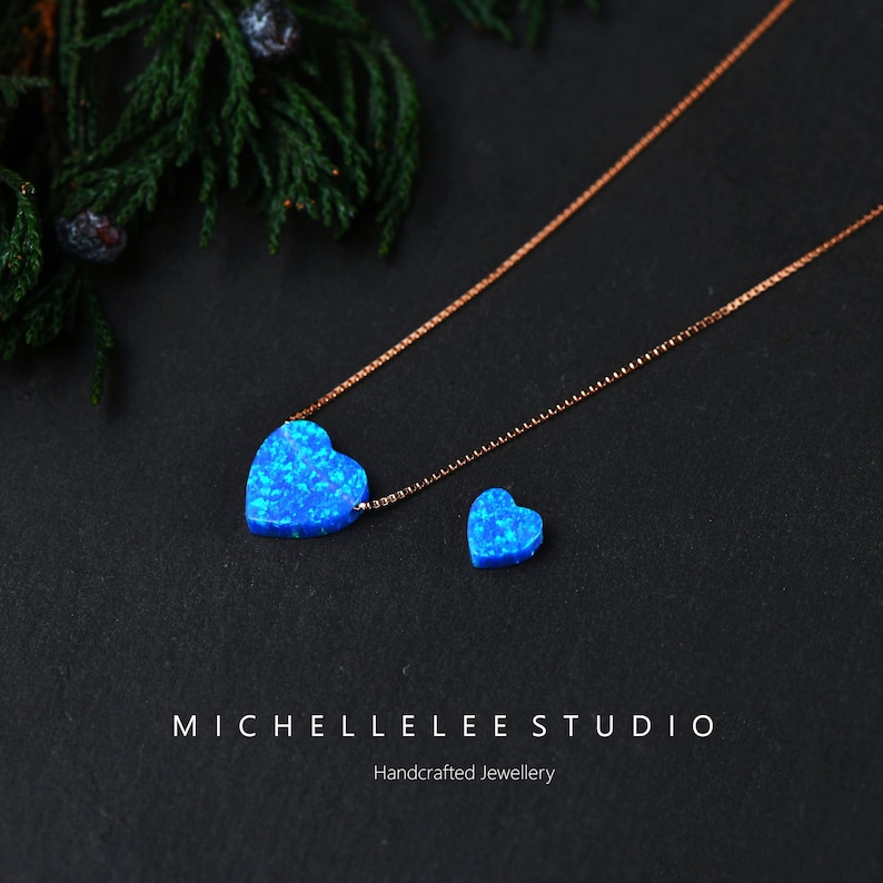 K&ouml;nnte beinhalten: Halskette und Ohrring-Set mit blauen herzf&ouml;rmigen Anh&auml;ngern. Die Halskette hat eine goldfarbene Kette. Der Ohrring ist eine kleinere Version des Anh&auml;ngers. Der Schmuck wird auf dunklem Hintergrund mit Gr&uuml;nzeug pr&auml;sentiert. Der Markenname ist sichtbar.