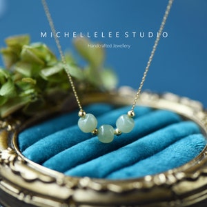 Peut inclure: Un collier délicat en chaîne dorée avec trois perles de jade vert clair. Le collier est présenté dans un écrin à anneaux en velours bleu. Le texte "MICHELLELEE STUDIO" et "Bijoux artisanaux" est visible au-dessus du collier.