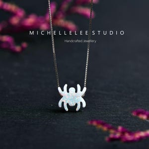 May include: A silver chain necklace with a white opal spider pendant. The pendant is a simple silhouette of a spider with eight legs. The necklace is on a black background with pink flowers.