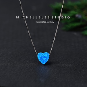 May include: A silver necklace featuring a heart-shaped, blue opal pendant. The pendant, a vibrant blue, hangs from a delicate chain. The necklace is set against a dark backdrop. The text "MICHELLELEE STUDIO Handcrafted Jewellery" is visible in the background.
