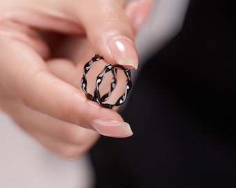 Puede incluir: Un par de pendientes de aro negros con un diseño trenzado y texturizado. Los pendientes presentan una superficie metálica continua y ondulada que crea un efecto de espiral. El texto MICHELLELEE STUDIO Handcrafted Jewellery es visible abajo.
