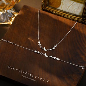 May include: A silver necklace and bracelet set featuring crescent moon and circular accents. The jewelry is displayed on a dark wooden surface. The brand name "MICHELLELEE STUDIO" is visible.