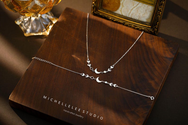 May include: A silver necklace and bracelet set featuring crescent moon and circular accents. The jewelry is displayed on a dark wooden surface. The brand name "MICHELLELEE STUDIO" is visible.