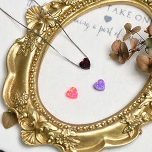 May include: A gold-coloured ornate frame surrounds a necklace with a dark red heart pendant and two heart-shaped gemstones. One gemstone is pink, the other is purple. Dried flowers are in the upper right corner.