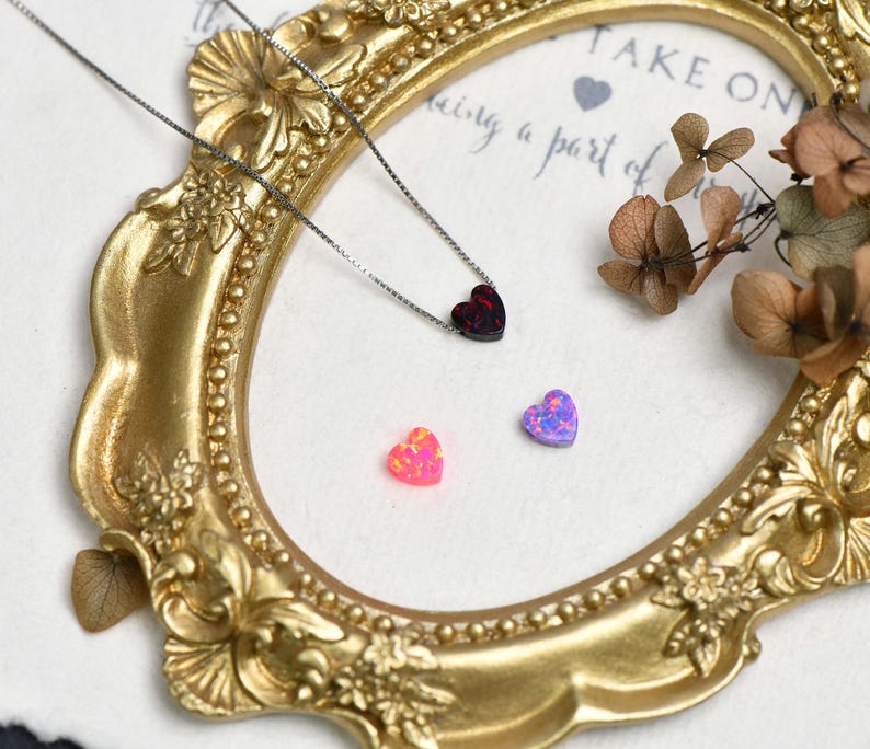 May include: A gold-coloured ornate frame surrounds a necklace with a dark red heart pendant and two heart-shaped gemstones. One gemstone is pink, the other is purple. Dried flowers are in the upper right corner.