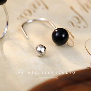 May include: Close-up of a pair of silver earrings with a black bead and a small silver bead. The earrings are on a cream-colored surface with the text "MICHELLELEE STUDIO" and "Handcrafted Jewellery".