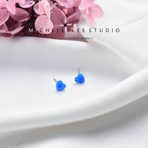 K&ouml;nnte beinhalten: Zwei blaue herzf&ouml;rmige Ohrstecker mit silbernen Stiften. Die Ohrringe bestehen aus einem schimmernden blauen Material. Der Text "MICHELLELEE STUDIO Handcrafted Jewellery" ist ebenfalls im Bild enthalten.