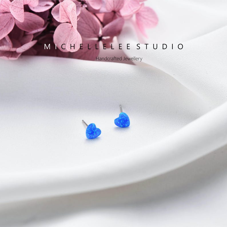 K&ouml;nnte beinhalten: Zwei blaue herzf&ouml;rmige Ohrstecker mit silbernen Stiften. Die Ohrringe bestehen aus einem schimmernden blauen Material. Der Text "MICHELLELEE STUDIO Handcrafted Jewellery" ist ebenfalls im Bild enthalten.