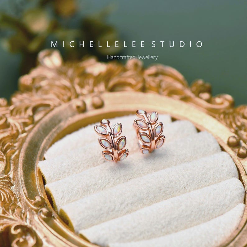 Puede incluir: Un par de pendientes de aro en oro rosa con dise&ntilde;o de hojas y peque&ntilde;as piedras ovaladas iridiscentes. Los pendientes se presentan sobre un inserto de terciopelo blanco en un marco dorado decorativo con la etiqueta MICHELLELEE STUDIO.