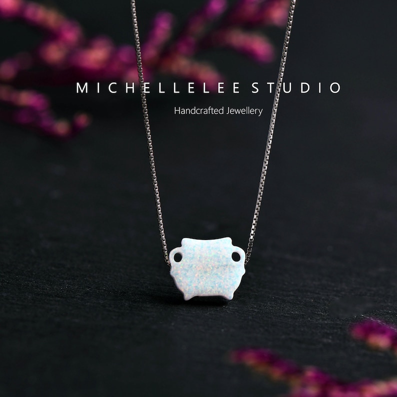 May include: A delicate silver chain necklace with a small, white opal pendant in the shape of a rounded rectangle. The pendant has a slightly textured surface and two small holes on either side. The necklace is photographed against a dark background with blurred purple flowers.