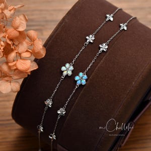 May include: Two delicate silver chain bracelets with flower-shaped opal pendants. The opal flowers have a white and blue iridescent color. The bracelets are displayed on a brown surface, with dried flowers in the background.