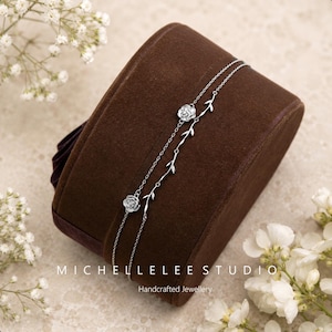 Könnte beinhalten: Ein silberfarbenes Doppelarmband mit zwei kleinen Rosenanhängern an einer Kette und einem zarten, rankenartigen Blattmuster an der anderen. Der Text MICHELLELEE STUDIO Handcrafted Jewellery ist unten im Bild aufgedruckt.