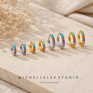 May include: A collection of small hoop earrings in silver and gold, each adorned with a row of colorful gemstones. The earrings are arranged on a neutral surface, showcasing their vibrant rainbow hues. The image includes the text "MICHELLELEE STUDIO Handcrafted Jewellery."