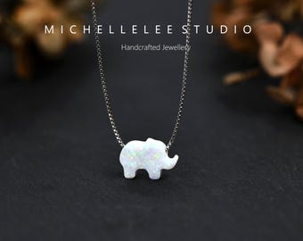 May include: A silver necklace featuring a small, white opal elephant pendant. The pendant has a textured surface and is suspended from a delicate chain. The background is dark, with the text "MICHELLELEE STUDIO Handcrafted Jewellery".