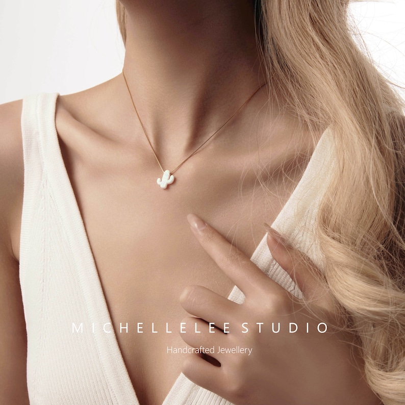 Op de afbeelding: Een delicate gouden ketting met een witte keramische cactus hanger. De ketting wordt gedragen door een model met lang blond haar. De tekst "Michelle Lee Studio" en "Handgemaakt juweel" is zichtbaar onder de hand van het model.