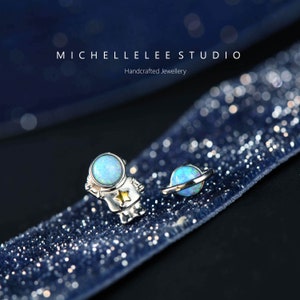 May include: Silver astronaut and planet stud earrings with blue opal stones. The astronaut has a yellow star on its chest. The earrings are on a dark blue, glittery surface. The text "MICHELLELEE STUDIO Handcrafted Jewellery" is at the top.