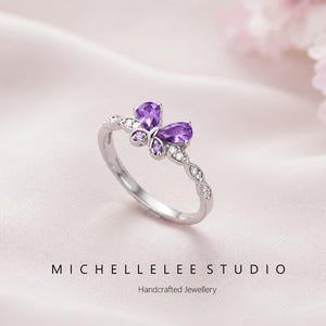 May include: A silver ring with a butterfly design, featuring pear-shaped purple gemstones and smaller purple and clear crystal accents. The ring band has a leaf-like pattern. The text "MICHELLELEE STUDIO Handcrafted Jewellery" is visible.
