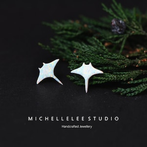 Op de afbeelding: Een paar witte opaal roggen oorknopjes. De oorbellen staan tegen een donkere achtergrond met groen gebladerte. De oorbellen zijn handgemaakte sieraden van MICHELLELEE STUDIO.