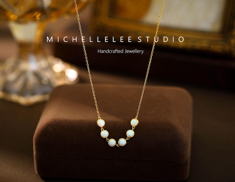 May include: A delicate gold necklace featuring a series of small, round, white gemstones. The necklace is displayed on a brown velvet surface. The text "MICHELLELEE STUDIO Handcrafted Jewellery" is visible.