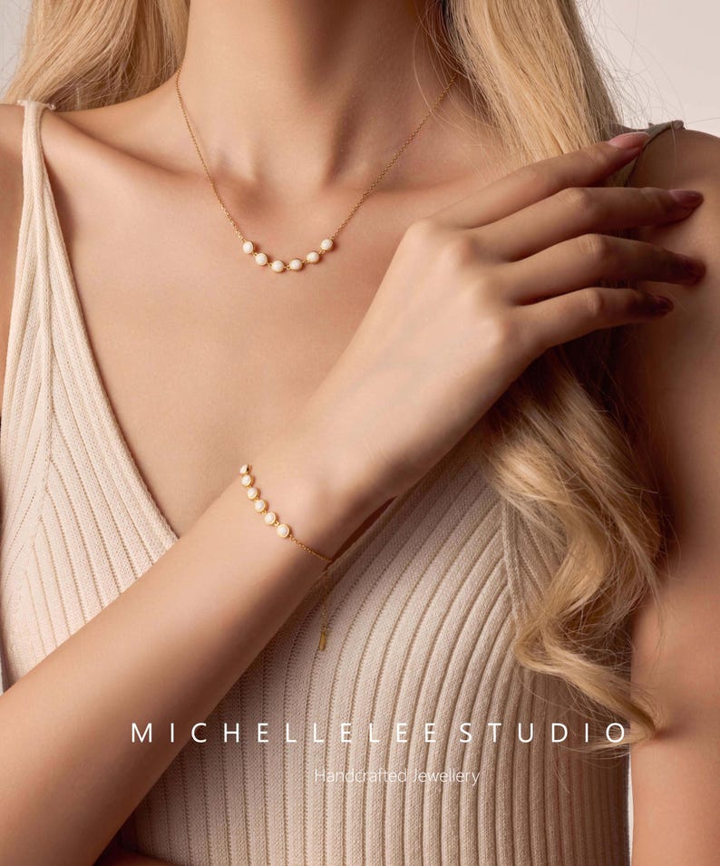 May include: A gold necklace and bracelet set featuring a series of small, white pearls. The necklace is a delicate design, and the bracelet is adjustable. The image also displays the text "MICHELLELEE STUDIO Handcrafted Jewellery".