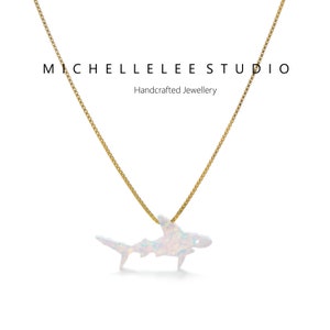 May include: A gold chain necklace with a white opal shark pendant. The pendant is a detailed silhouette of a shark with a shimmering opal finish. The necklace is a delicate piece of jewelry that would make a unique gift for a shark lover.