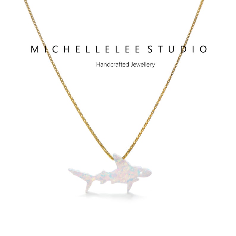 May include: A gold chain necklace with a white opal shark pendant. The pendant is a detailed silhouette of a shark with a shimmering opal finish. The necklace is a delicate piece of jewelry that would make a unique gift for a shark lover.