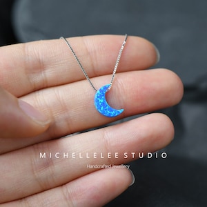 May include: A silver necklace featuring a crescent moon pendant with a blue opal-like design. The necklace is displayed on a hand, with the brand name "MICHELLELEE STUDIO" and the text "Handcrafted Jewellery" visible.