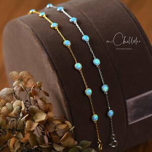 Pode incluir: Duas pulseiras delicadas com pedras redondas de opala azul claro. Uma pulseira &eacute; dourada, a outra prateada. As pulseiras s&atilde;o exibidas em uma superf&iacute;cie de veludo marrom, com flores secas em primeiro plano. A marca "mi Chellelee" &eacute; vis&iacute;vel.