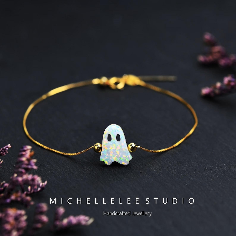 Pu&ograve; includere: Un bracciale a catena dorata con un ciondolo centrale a forma di fantasma in opale bianco iridescente con due occhi circolari neri. Il ciondolo &egrave; affiancato da due piccole perline dorate. Il testo MICHELLELEE STUDIO Handcrafted Jewellery &egrave; visibile.