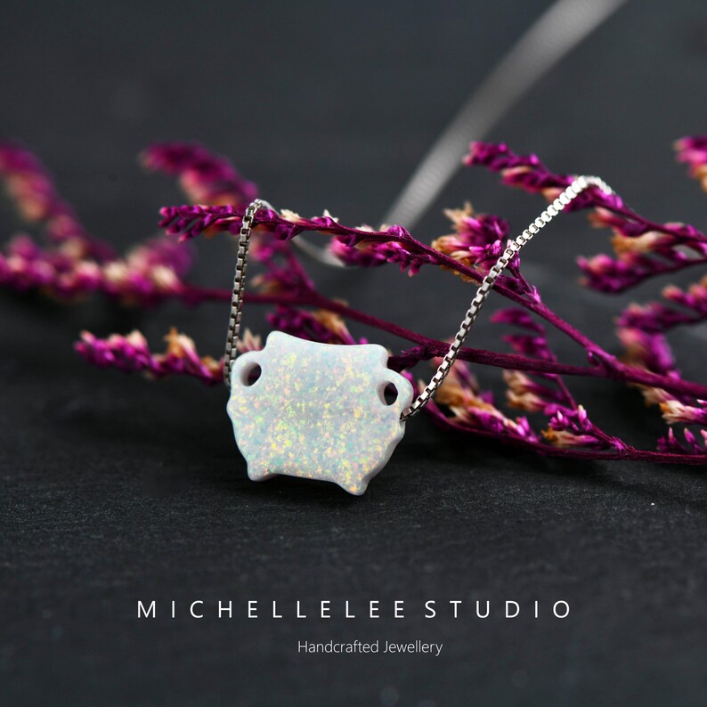May include: A delicate silver chain necklace with a white opal pendant shaped like a small vase. The pendant has a sparkly, iridescent finish. The necklace is photographed on a background of pink flowers.