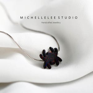 May include: A silver chain necklace with a black opal spider pendant. The spider is a dark red color with iridescent sparkles. The necklace is on a white background.