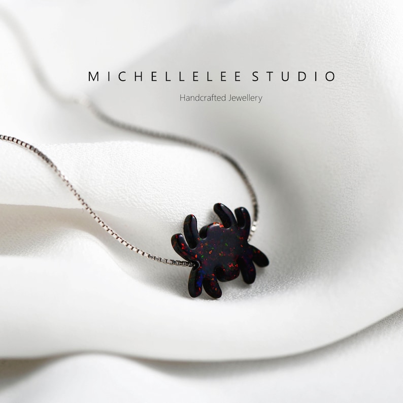 May include: A silver chain necklace with a black opal spider pendant. The spider is a dark red color with iridescent sparkles. The necklace is on a white background.