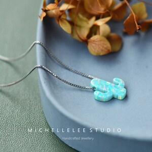 Op de afbeelding: Een zilveren ketting met een blauwe opaal cactus hanger. De hanger is lichtblauw met witte en groene accenten. De ketting ligt op een blauw bord met gedroogde bloemen op de achtergrond. De tekst "MICHELLE LEE STUDIO" en "Handgemaakt juweel" is zichtbaar onderaan de afbeelding.