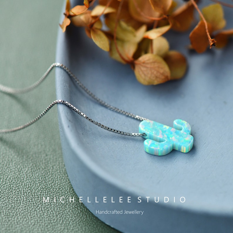 Op de afbeelding: Een zilveren ketting met een blauwe opaal cactus hanger. De hanger is lichtblauw met witte en groene accenten. De ketting ligt op een blauw bord met gedroogde bloemen op de achtergrond. De tekst "MICHELLE LEE STUDIO" en "Handgemaakt juweel" is zichtbaar onderaan de afbeelding.