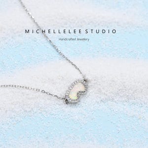 May include: A delicate silver necklace featuring a pendant with a white opal center and a halo of small, clear crystals. The pendant is shaped like a stylized heart or teardrop. The necklace is displayed on a light blue and white textured surface. Text on the image reads "MICHELLELEE STUDIO Handcrafted Jewellery".