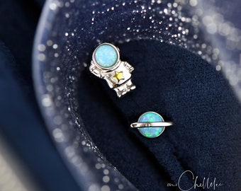 Puede incluir: Un par de pendientes de plata. Uno es un astronauta con cabeza de ópalo azul y estrella amarilla. El otro es un planeta con centro de ópalo azul y anillo plateado. Fondo azul oscuro.