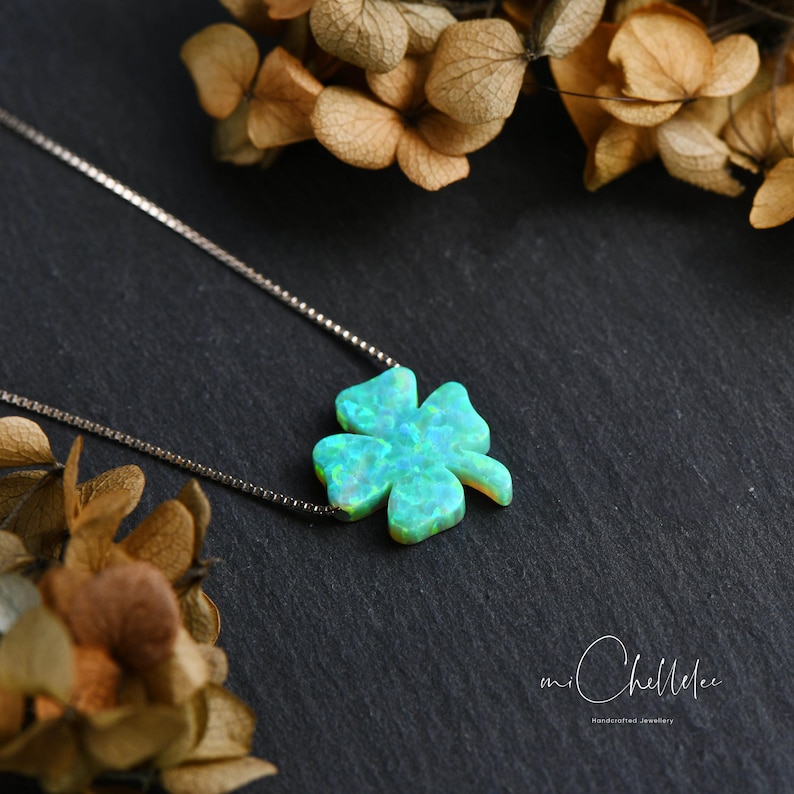 May include: A silver necklace with a four-leaf clover pendant in shimmering opalescent green and blue. The pendant is set against a dark backdrop with dried flowers, creating a striking contrast. The necklace is a delicate piece of jewellery.