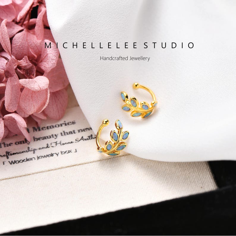 Puede incluir: Dos pendientes tipo ear cuff dorados con dise&ntilde;o de hoja e incrustaciones ovaladas de color azul claro. El texto MICHELLELEE STUDIO Handcrafted Jewellery aparece sobre las joyas, situadas junto a p&eacute;talos de hortensia rosas.