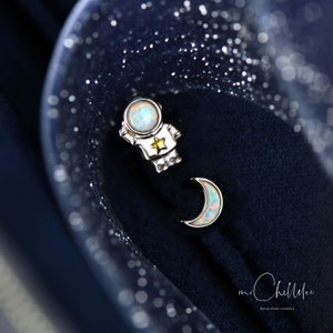 May include: A pair of silver earrings featuring an astronaut and a crescent moon. Both charms are adorned with iridescent opal stones.