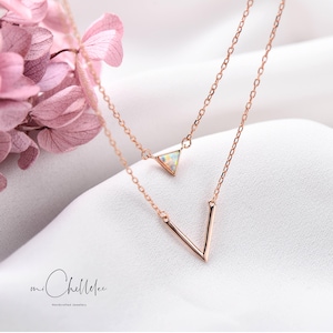 May include: A rose gold layered necklace featuring a triangle pendant with an iridescent stone and a V-shaped pendant. The necklace is displayed on a white surface with pink flowers. The brand name "mi Chellelee" is at the bottom.