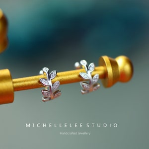 Puede incluir: Un par de pendientes de aro de plata con forma de hoja, cada uno con peque&ntilde;as piedras transparentes incrustadas a lo largo del dise&ntilde;o de rama. El texto MICHELLELEE STUDIO Handcrafted Jewellery aparece impreso en la parte inferior.