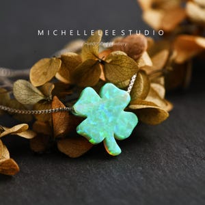 May include: A silver necklace with a four-leaf clover pendant. The pendant has an iridescent, light green finish with blue and orange hues. The necklace is from Michelle Lee Studio, which is described as handcrafted jewellery.